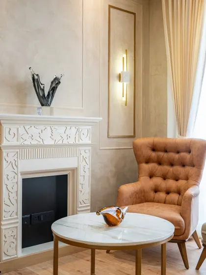 This cozy corner exudes timeless elegance with its beautifully carved fireplace and a comfortable tufted armchair. The striking glass sculptures and marble-top table add a touch of modern luxury to the refined space.