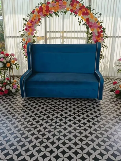 A simple and elegant floral arch for a small event or photo booth. This setup features a blue sofa and two floral stands, perfect for a daytime function.
