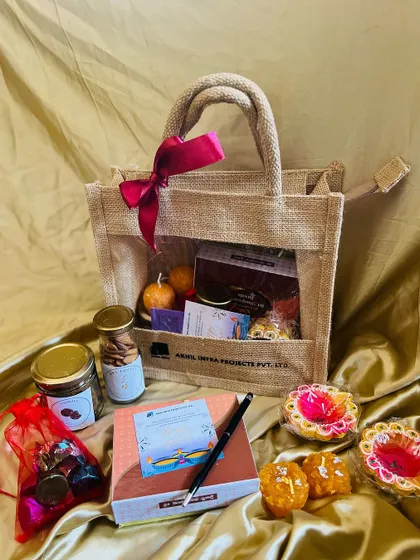 A jute bag hamper displayed with some of its contents outside the bag, including a notebook, pen, and festive diyas.