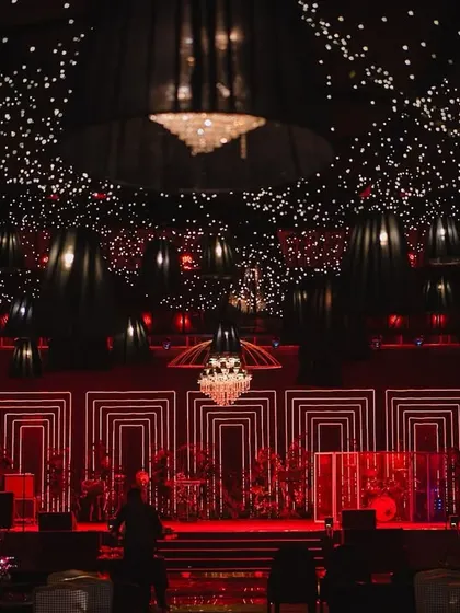 A dazzling setup for a sangeet night, where a ceiling of countless lights and hanging lamps creates a celestial glow. The stage, lit in dramatic red with geometric patterns, sets the scene for an unforgettable evening of performances and celebration.