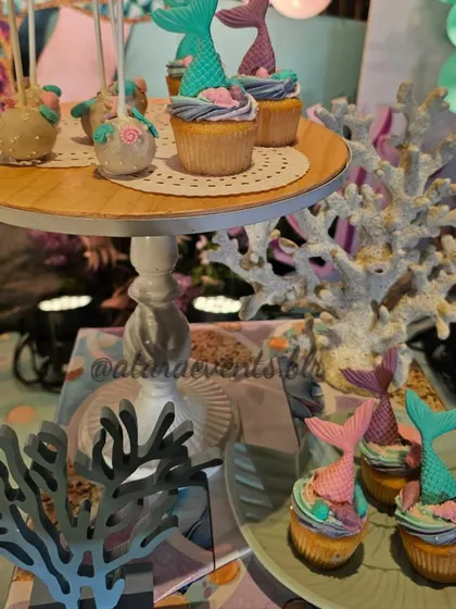 A dessert table from a mermaid-themed party, featuring cupcakes with colorful mermaid-tail toppers. The treats are displayed on a tiered stand, surrounded by coral and other sea-inspired decorations.