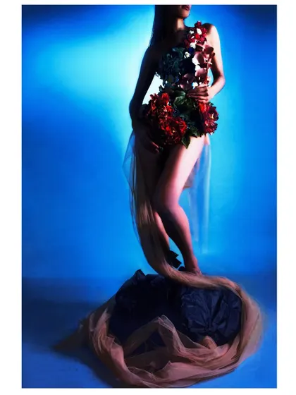 A silhouette shot against a deep blue background, focusing on the form of the floral garment and the model's pose. The image is dramatic and sculptural, highlighting the artistic styling.
