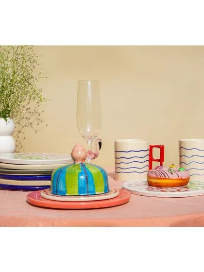 A still life of the colorful tableware set, including a striped butter dish, wavy-handled mugs, and a flamingo-stemmed glass. The composition is playful and modern.