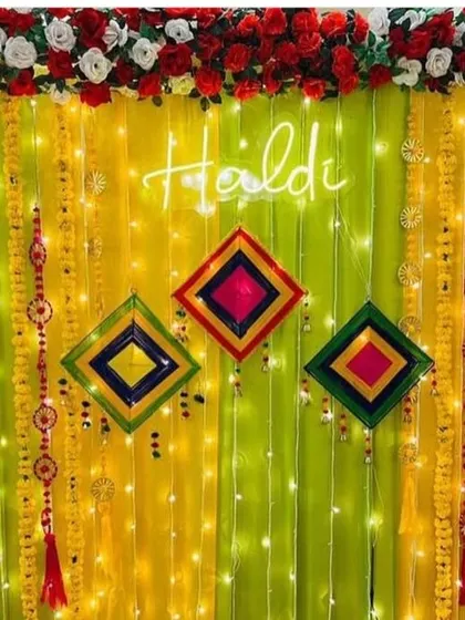 A close-up of a Haldi backdrop with a neon sign, showing the details of the hanging marigold garlands and colorful kite props against a lime green drape.