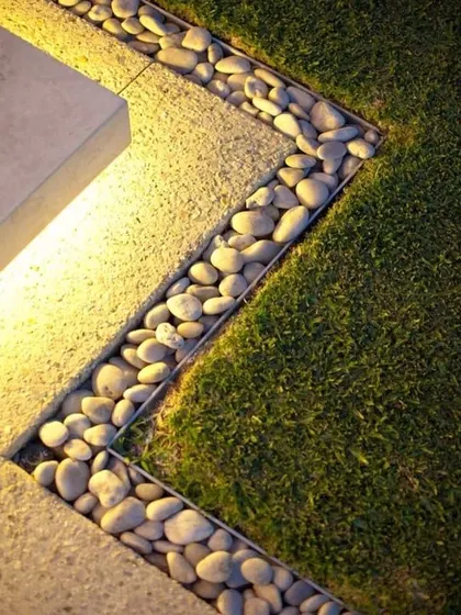 A close-up detail showing a clean border between a lawn and a paved area, filled with smooth river pebbles and accented with lighting. This is a modern and stylish finishing touch.