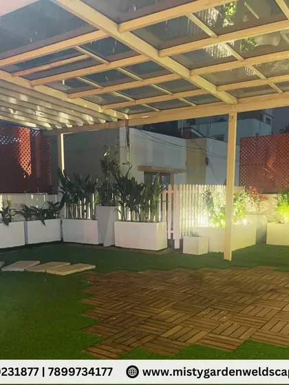 A night view of a terrace garden in Banashankari, featuring a glass pergola, artificial grass, and FRP planters, designed as a peaceful family gathering space.