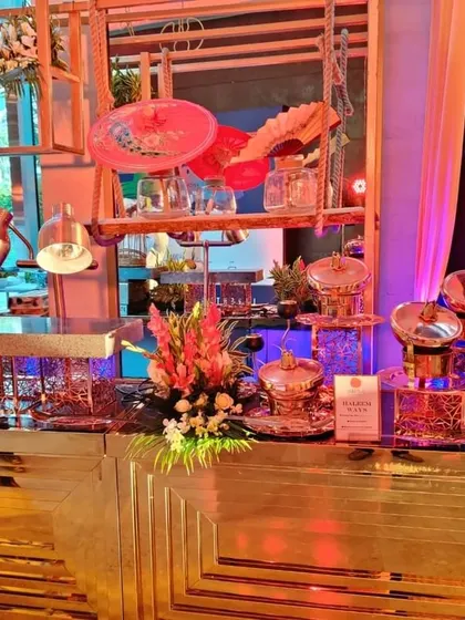 An elegant, modern food counter with gleaming copper chafing dishes, floral decor, and integrated heat lamps. This setup, with a chef visible in the background, showcases our capacity for large-scale, stylish event catering.