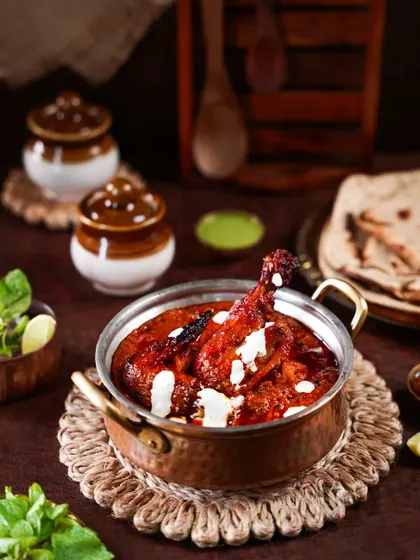 A delicious chicken curry in a copper pot, garnished with cream. The composition with traditional pickle jars and flatbread in the background tells a story of a complete, wholesome Indian meal.