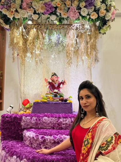 This Ganpati setup combines a rich purple floral base with a canopy of pastel flowers and rustic dried grasses. The "Ganpati Bappa Morya" sign adds a beautiful, personalized touch to the decor.
