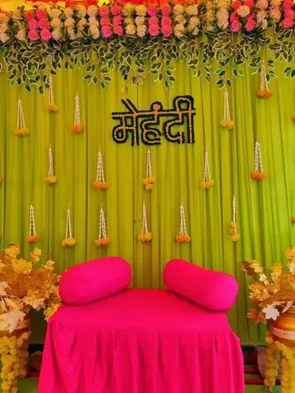 A simple yet beautiful Mehendi backdrop with a light green drape, hanging floral strings, and a 'Mehendi' sign. The bright pink seating provides a lovely color contrast.