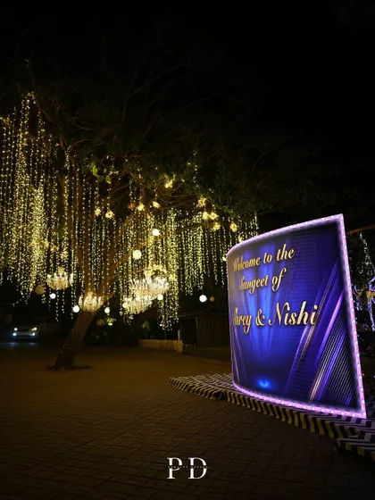 A modern, curved LED welcome sign for a 'Starry Night' Sangeet, set against trees adorned with hanging chandeliers and fairy lights.