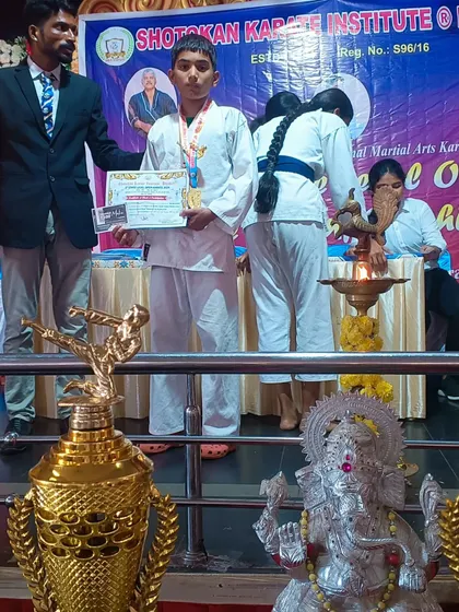 A young student receives his medal and certificate at the Shotokan Karate Institute's state-level championship. The trophies in the foreground show the scale of the competition.