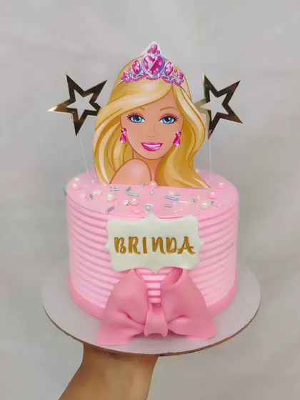A classic Barbie theme cake with a pink striped buttercream finish. The design is completed with a large fondant bow and a Barbie silhouette topper.