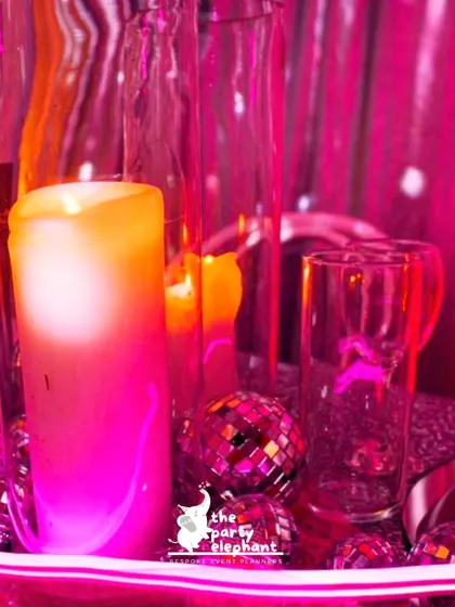 A close-up of a table centerpiece at the disco party, showing a candle and mini disco balls bathed in a vibrant pink light, creating a moody and glamorous feel.
