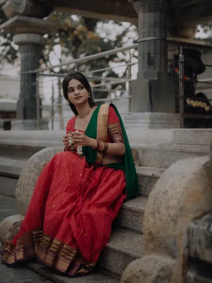 A serene moment captured at a temple. The model, dressed in a traditional red and green half saree, sits on ancient stone steps, creating a timeless and culturally rich image.