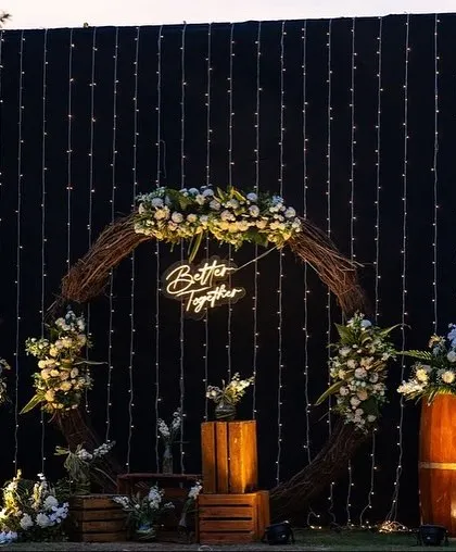 A rustic-chic photo backdrop for a pre-wedding function. A "Better Together" neon sign is placed within a wreath of twigs and white flowers, against a wall of fairy lights.