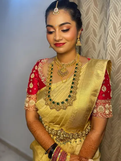 A full view of the bride's muhurtham look. The makeup is subtle yet defined, with a focus on a champagne gold eyeshadow that complements her golden saree and emerald jewelry.