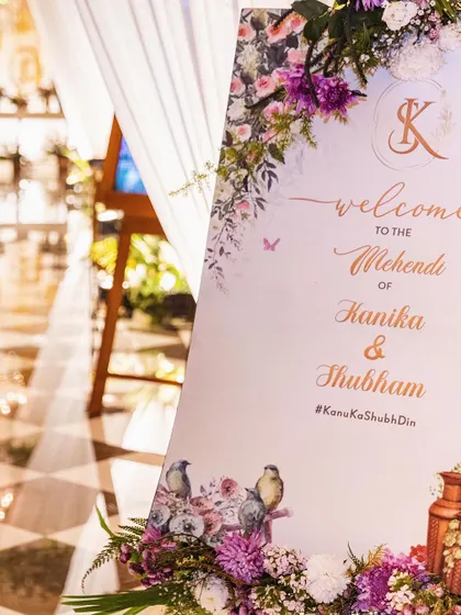 A beautifully designed welcome sign for a Mehendi, featuring a delicate floral and bird motif with purple and pink flowers that match the event's color scheme.