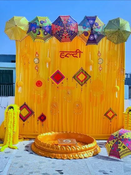 We design vibrant and traditional setups for pre-wedding ceremonies. This outdoor Haldi decoration includes a bright yellow backdrop with the word 'Haldi' in Hindi, decorated with colorful Rajasthani umbrellas, marigold garlands, and traditional hangings.