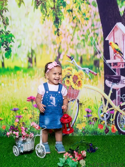 A toddler's happy laugh captured in a vibrant outdoor park-themed studio set. The bicycle, birdhouse, and colorful flowers create a joyful and lively scene.
