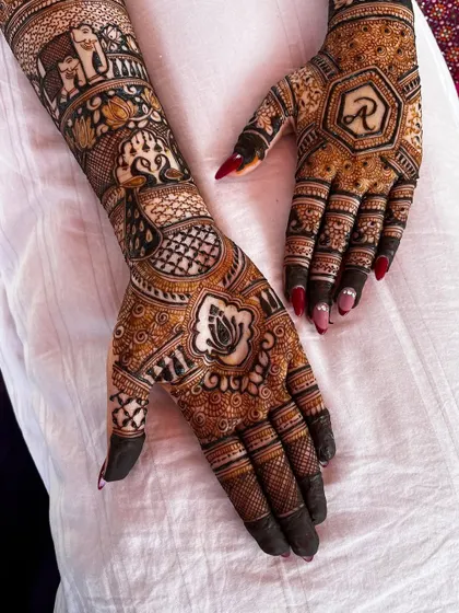 A close-up of a bridal mehendi featuring a lotus motif and the groom's initial 'R' inside a hexagon. The surrounding design is filled with intricate details and shading.