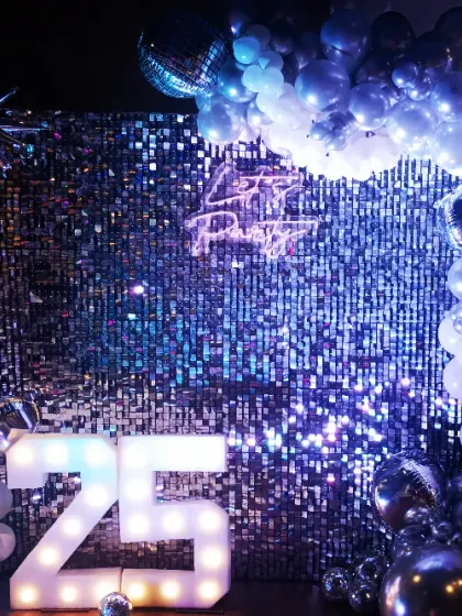 Celebrating a 25th anniversary with a touch of disco fever. This setup includes a silver sequin wall, a "Let's Party" neon sign, and a black and silver balloon garland with starburst accents.