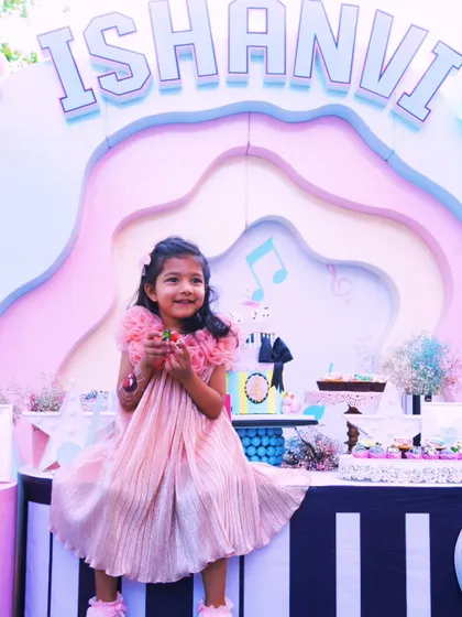 A beautiful shot of the birthday girl, Ishanvi, sitting on her piano-themed dessert table, surrounded by pastel colors and musical decor. A moment of pure sweetness.