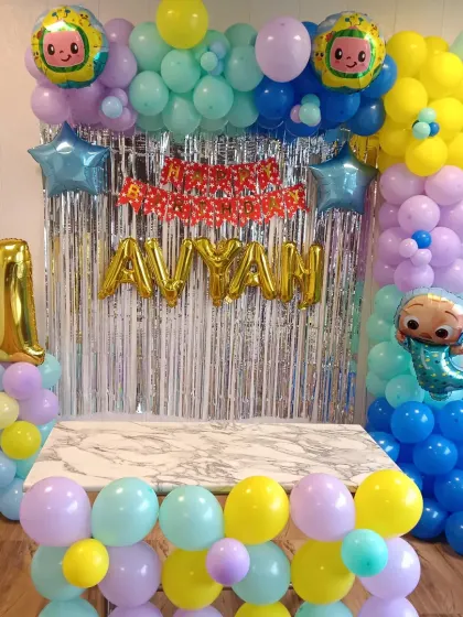 A bright and cheerful Cocomelon theme for a first birthday. This decoration uses a silver tinsel curtain backdrop, a colorful balloon frame, Cocomelon character foil balloons, and gold foil balloons for the name "AVYAN" and the number '1'.