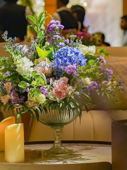 A beautiful floral arrangement in a glass bowl, set on a modern coffee table. The mix of blue hydrangeas, pink roses, and other blooms creates a soft and romantic look.