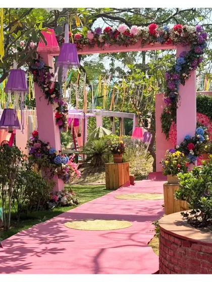 A beautiful and colorful entrance for a Haldi ceremony. The pink archway is adorned with multi-colored flowers and hanging lamps, creating a whimsical and inviting pathway.