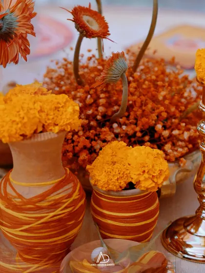 A close-up of a traditional table centerpiece, featuring orange marigolds and gerberas in rustic, thread-wrapped terracotta pots.