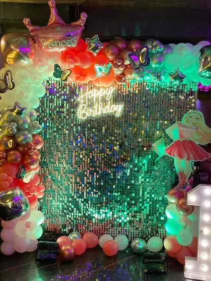 A glamorous mermaid party with a sparkling sequin backdrop. The decoration includes a multi-color balloon arch, a mermaid cutout, and a light-up number '1', all lit with green mood lighting.