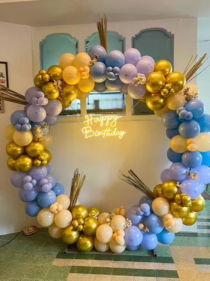 A chic and modern circular balloon arch using a pastel color palette of purple, blue, and yellow, with pops of metallic gold. The "Happy Birthday" neon sign and decorative dried grass give it a unique, bohemian touch.