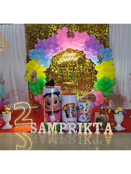 A bright and colorful Cocomelon theme for a second birthday, set against a gold sequin wall. The setup includes character cutouts, a rainbow balloon arch, and a light-up number '2'.