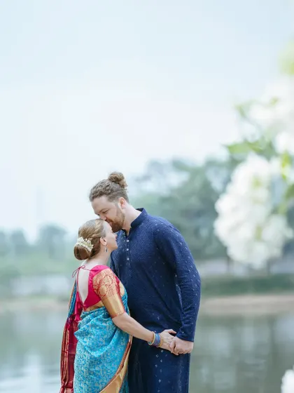 A tender moment between a multicultural couple by the water, the groom kissing the bride's forehead. Love knows no boundaries.