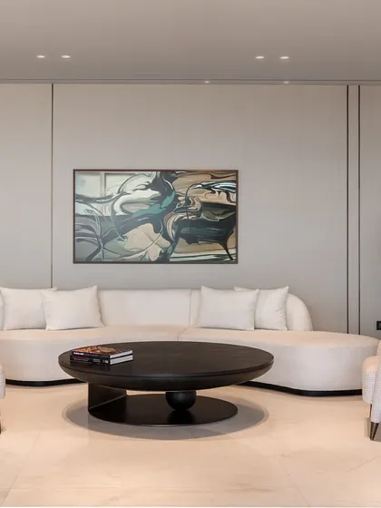 A seating area featuring a custom curved sofa and matching chairs in a neutral tone. The dark oak veneer coffee table with an antique brass detail provides a grounding element. The backdrop of wallpaper and veneer paneling adds a layer of subtle sophistication.