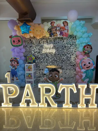 A Cocomelon theme party with a silver shimmer wall, character cutouts, and large light-up marquee letters spelling the birthday boy's name.