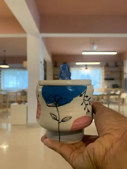 A lovely lidded jar, hand-painted with delicate flowers and topped with a little Buddha figure. This student's work is a beautiful blend of artistic skill and personal expression.