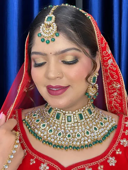 A classic bridal look with a red lehenga and green kundan jewelry. The eye makeup is a soft gold shimmer, paired with a deep red lip for a timeless and elegant finish.