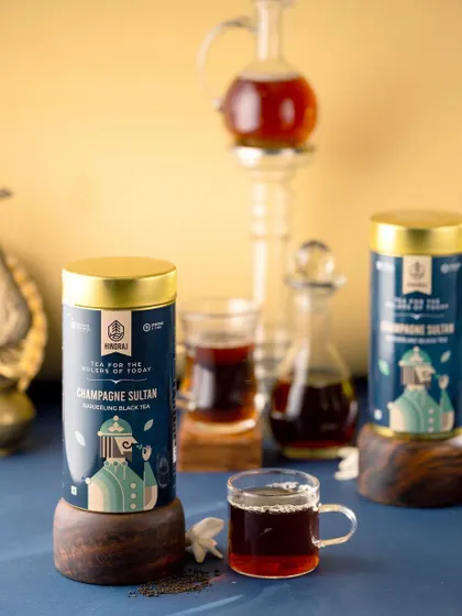 A regal and sophisticated shot of Hindraj's Champagne Sultan tea. The product is styled with antique-looking glassware and wooden stands against a royal blue background, giving it a premium and luxurious feel.