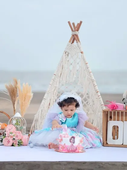A boho-chic first birthday cake smash on the beach, featuring a macrame teepee and pampas grass against the ocean backdrop.