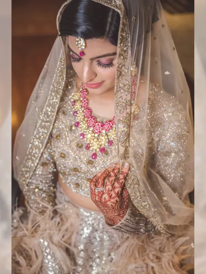 A classic bridal portrait, with the bride looking down, veiled and elegant. The focus is on her intricate, feathered lehenga and stunning ruby necklace.