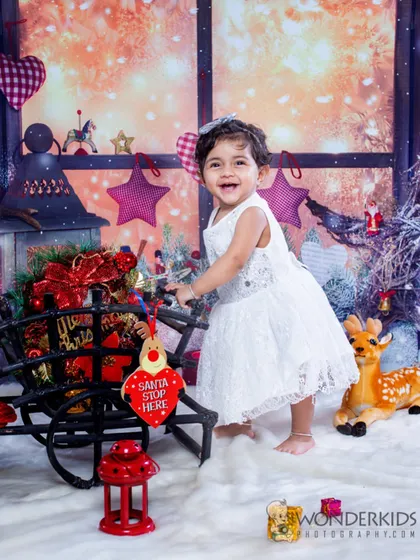 A happy toddler in a white dress enjoys a magical Christmas-themed setup, complete with a sleigh, reindeer, and a snowy window view. Her bright smile perfectly captures the holiday spirit.