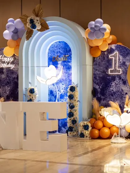 A luxurious hot air balloon theme for a first birthday. The setup features large "ONE" marquee letters, glowing floral lamps, and a stunning blue marble-effect backdrop, creating a dreamy and elegant atmosphere.