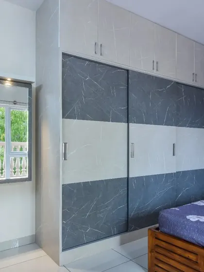 A bedroom with a modern sliding wardrobe featuring a marble-finish pattern. The compact dressing mirror mounted on the wall is a smart, space-saving solution.