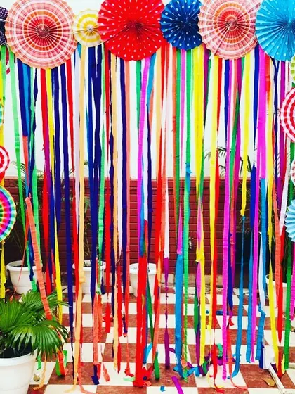 A colorful and traditional backdrop designed for a Holi celebration. This decoration uses vibrant paper fans and long, flowing streamers to create a festive and joyful photo background for an outdoor or daytime event.