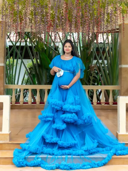 A grand and beautiful portrait of a mom-to-be in a stunning blue ruffled gown. She is holding a small pair of baby shoes, standing on a veranda with hanging flowers.