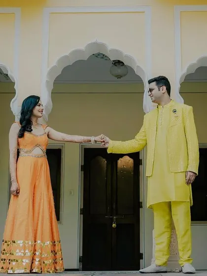 A sweet moment during a haldi celebration at a palace. The couple holds hands, their bright yellow and orange outfits standing out against the beautiful architecture.