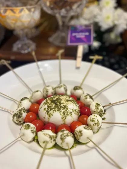A modern presentation of a Caprese salad. Cherry tomatoes and mozzarella balls are skewered and arranged around a ball of fresh burrata, drizzled with pesto.