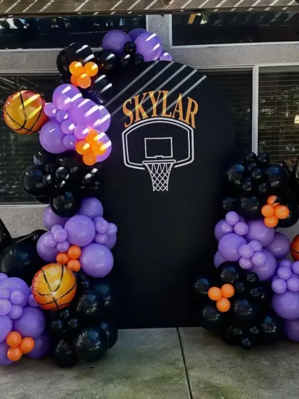A dynamic basketball-themed party with a custom backdrop featuring a hoop, and a balloon arrangement in purple, black, and orange.